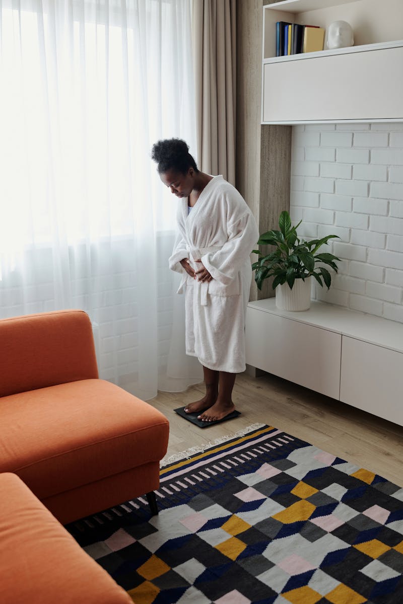 African American woman in a bathrobe checking her weight on a scale in a modern living room.