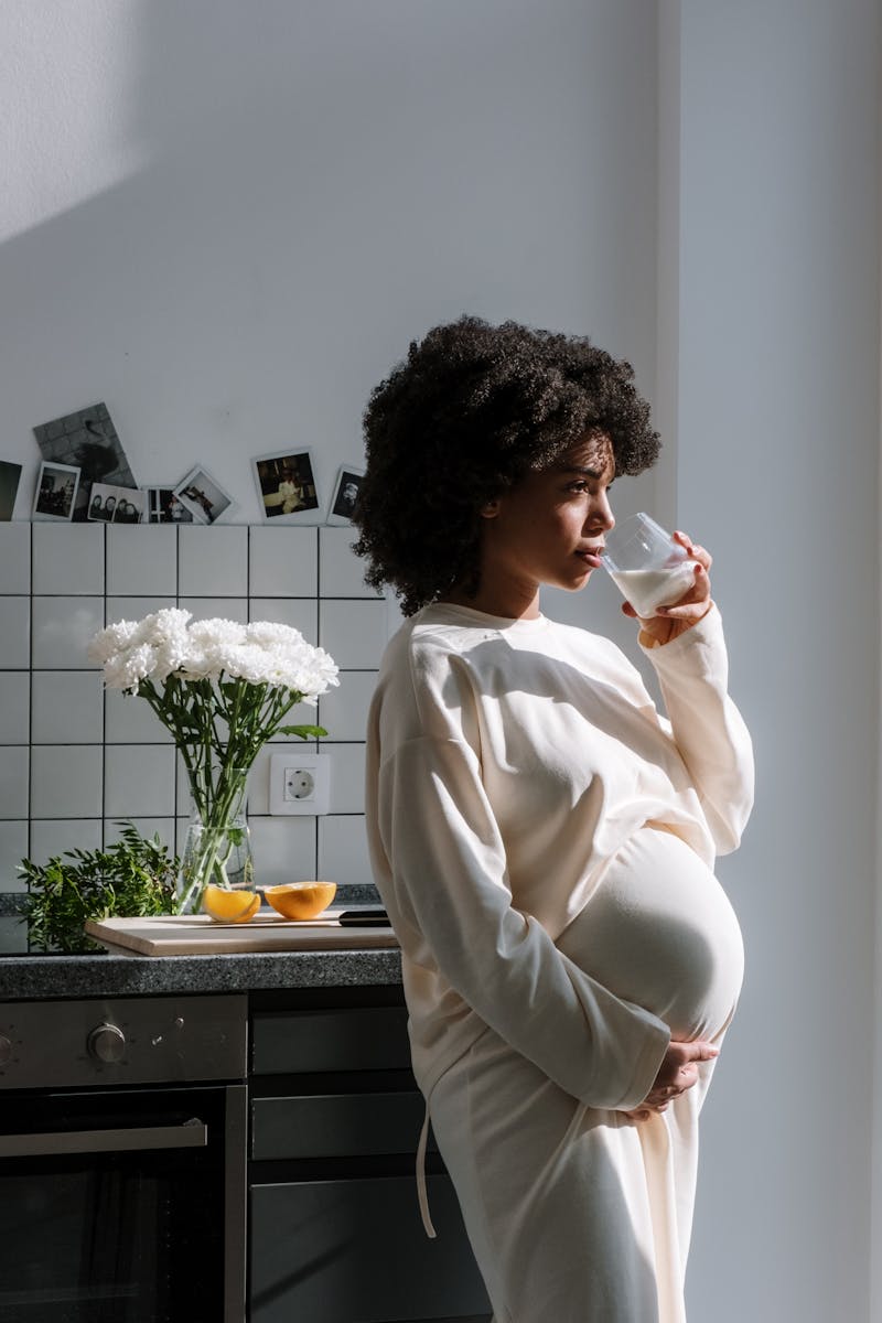 Pregnant woman drinking milk in a sunlit kitchen, surrounded by flowers and fruit.